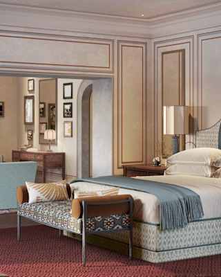 A bed with patterned upholstery, blue throw and bench, seen in the airy Limonaia Suite, with pale décor and russet carpet.