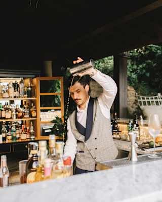 Waistcoated barman pouring a cocktail from a shaker in a bar surrounded by liquor bottles