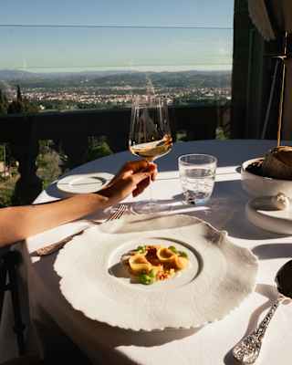 A woman's hand holds a glass of pale rosé next to an elegant, wide-rimmed dish of tortellini, at a table with sublime city views.