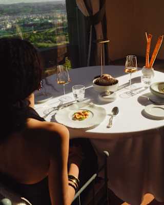 View over a female diner's shoulder of a table at Antesi restaurant, with an elegant plate of pasta, wine, bread and views.