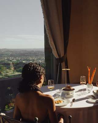A man and woman dine at a round table by a large window with a scenic city view. Sunlight streams in, illuminating their meals and wine glasses on the table.