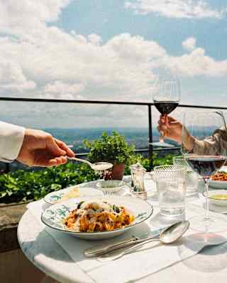 A waiter sprinkles parmesan onto an unctuous pasta dish as a guest opposite raises a glass of red wine, at an al fresco table.