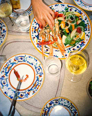 A diner picks from a dish of langoustines and salad at a lunch table with wine and Deruta crockery with Florentine patterns.