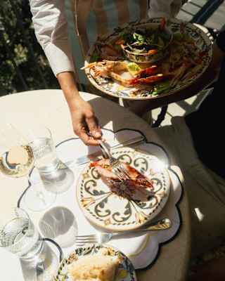 A waiter places seafood, including crab legs, onto a decorative plate at an outdoor table set with water glasses, bread, and salad. Sunlight casts shadows across the scene.