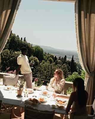 Tilted image of a man standing by a table on a covered terrace with tied drapes, where guests enjoy lunch with sublime views.