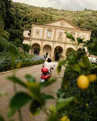 A man on a red vespa drives along the road towards Villa San Michele, in an angled shot taken from behind a lemon tree.