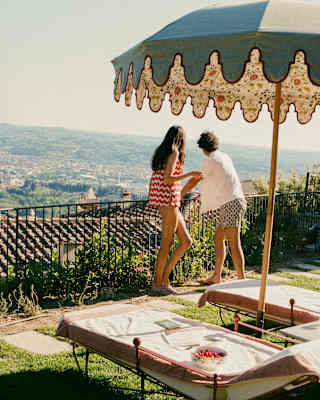 A woman in a red and white top and a male friend gaze at views from the terrace, seen over loungers and a pretty floral parasol.