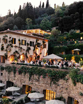 Guests mingle on the terrace as warm window and garden lights provide a magical evening atmosphere, seen across a courtyard.