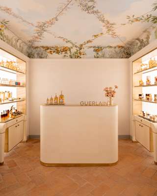 Backlit product shelves flank a white spa reception desk with a row of bottles, a Guerlain sign and a vase of pink blooms.