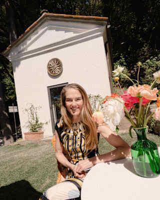 J.J Martin, with long blonde hair, smiles to camera, seated at a table with a flower vase in gardens by the energy chapel.