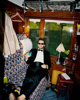 A man in dark glasses and a tuxedo holds a blue drink as he reclines on the banquette of a historic wood-panelled cabin.