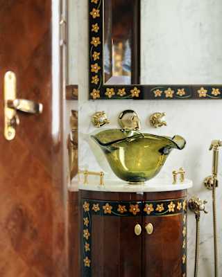 Beyond a polished bathroom door, a walnut unit with a marble top supports a green handblown glass sink in a design detail.