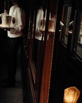 A steward in white jacket stands half in shadow in a carriage corridor holding a tray with a wine cooler and bottle of champagne.
