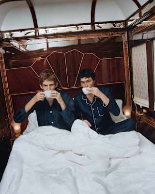 Two men in navy pyjamas sip coffee beneath the white duvet of their bed in the Prague Suite, photographed by Coco Capitán.