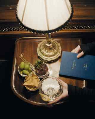 A guest's hands hold a blue book titled Writers on the Rails, and a drink, seen from above at a lamp-lit table with snacks.