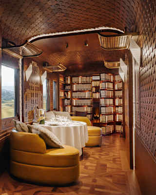 A lounge view glances a window-side daybed with book and binoculars into a calm space with yellow chairs, table and library.