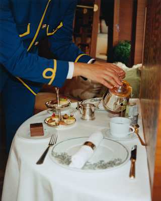 A train steward in a blue uniform with gold braid pours Afternoon Tea at a demilune table for two, seen from the neck down.