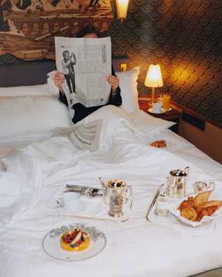 View from the foot of a bed, over breakfast trays to a guest, propped up on a pillow, reading a paper that covers their face.