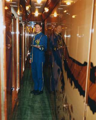 A smiling female train attendant in a blue uniform stands in a narrow, glossy train corridor, holding a drink on a tray.