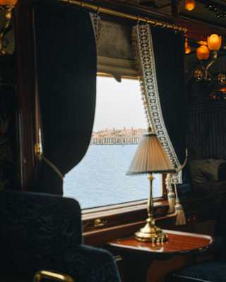 At a bar car window table with a lamp and velvet seats, hemmed blue curtains are hooked back to reveal wide water views.