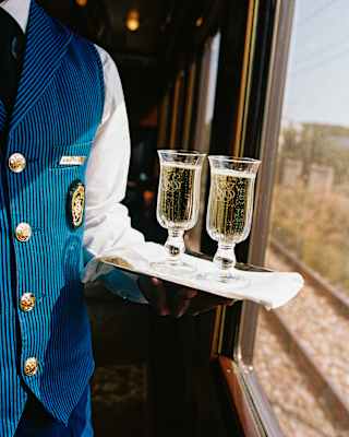A waiter in a blue and black-striped uniform, seen from the neck down, holds a tray with two glasses of Champagne by a window.