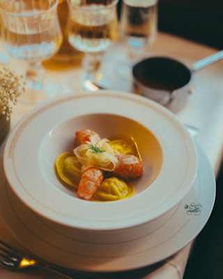 A white bowl with ravioli, topped with prawns and herbs, sits on a plate. Wine glasses and a cup are on the elegant, softly lit table in the background.