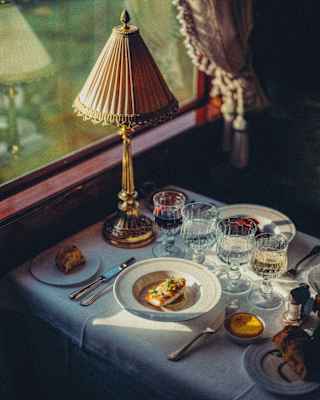 Sunlight falls onto a dining table with a plate of grilled white fish and another main, obscured by four crystal wineglasses.