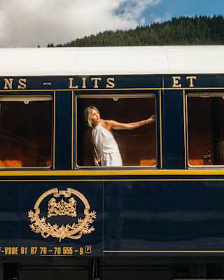 A blond woman in a white halter-neck dress stretches an arm across the train window as she peers out, seen from the platform.