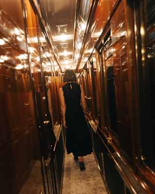 A female guest in a black dress walks down an interior, windowless corridor lit with bright ceiling lights, seen from behind.