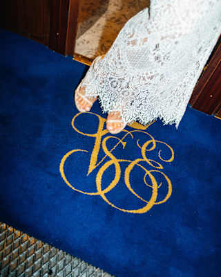 Seen from the waist down, a woman in a white lace dress and heels stands on a blue velvet carpet with the gold VSOE monogram.