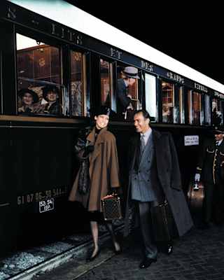 A couple dressed elegantly in coats and hat walk by the side of the carriage. Behind them gold-trimmed stewards carry bags