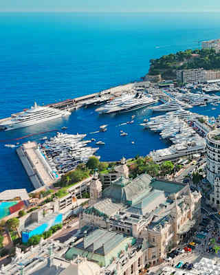 Aerial view of a marina filled with yachts and boats, surrounded by modern and historic buildings, with the sea and hills in the background.