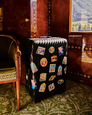 A black suitcase covered with pictorial travel stickers sits by an antique chair inside a luxury train suite below a window.