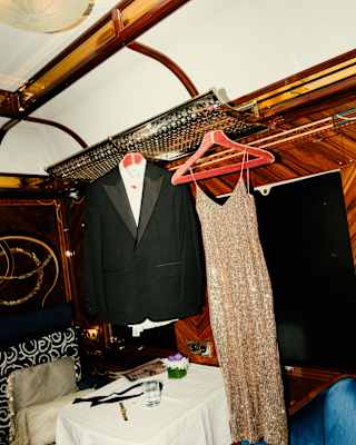 A sparkling gold evening dress and a tuxedo dangle from coat hangers above a table and a blue velvet chair in a cabin.