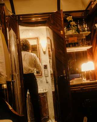 A man in black trousers and white shirt checks his tie in the bathroom mirror, seen from the warm-lit cabin interior behind.