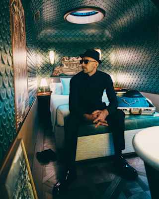 Artist JR, in black clothes, hat and shades sits on the end of a bed in the leather-tiled cabin, gazing at a marquetry panel.