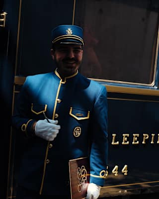 Chiaroscuro image of a train guard in a uniform of royal blue with gold piping and hat as he stands in a carriage doorway.