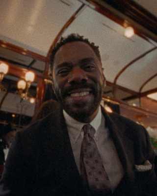 Colman Domingo, in a jacket, shirt and tie, smiles to camera in a low-angle image capturing interior carriage lights above.