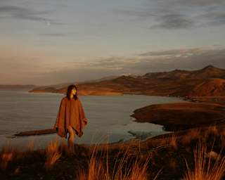 A woman in a brown wool poncho strides up an incline with a backdrop of hill chains and calm lake waters, seen at sunset.