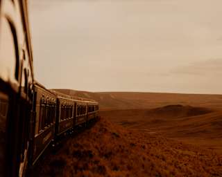 External view from the fifth carriage window capturing the train front forging through grasslands tinted deep-red by sunset.