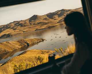 Angled image of a woman, in soft-focus, silhouetted against a window with crisp views across a lake edged with gold grasses.