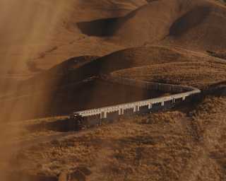 The Andean Explorer train journeys along a snaking track through undulating hills, seen from above through golden sun haze.