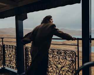 A guest in a long brown coat leans on the observation deck's filigree rails gazing out over gold grasslands and a blue lake.