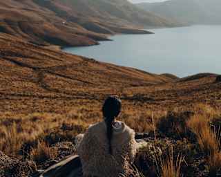 A woman with a long dark plait, wearing a wool shawl, sits on a blanket in grassland above Lake Saracocha, seen from behind.