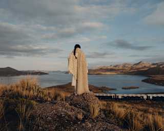 A woman wearing a white woollen shawl watches the train from a high rock as it passes Lake Saracocha below, seen from behind.