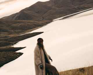 Tilted image of a woman in a long white coat and cap as she strides across a grassy hill overlooking Lake Saracocha.