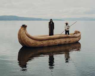 A figure in dark clothes stands in a banana-shaped vessel of bound totora reeds on Lake Titicaca, steered by a local boatman.