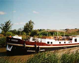 The Alouette sails the smooth brown waters of the canal in pleasant countryside beneath blue sky, seen over a grassy bank.