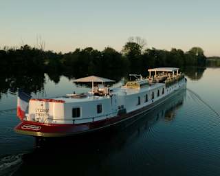 Soft sun brushes the canopy on the deck of Coquelicot as it glides along a wide, tree-lined waterway, seen from behind.
