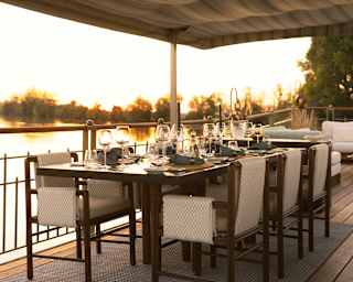 Beneath a canopy, the dining table on deck is laid with water and wineglasses, and blessed with golden sunset water views.